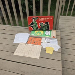 May include: A Strat-O-Matic Baseball board game set featuring a red box with green and black illustrations of baseball players, a green game board, orange square tokens, white paper score sheets, a fielding chart, and small game cards.