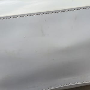 Op de afbeelding: Een gladde witte handtas met een glanzende afwerking en zichtbare lichtgrijze stiksels langs de horizontale naden. Het oppervlak van de tas vertoont subtiele gebruikssporen met vage donkere vlekken en kleine krasjes op het witte materiaal.