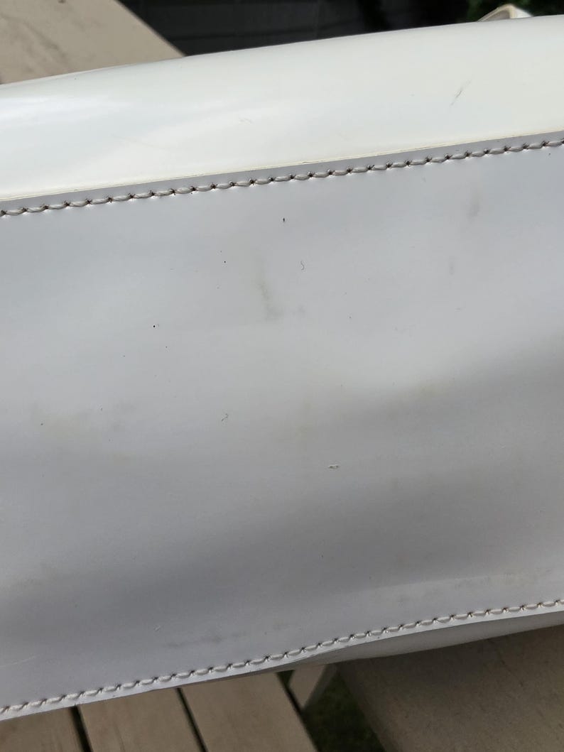 Op de afbeelding: Een gladde witte handtas met een glanzende afwerking en zichtbare lichtgrijze stiksels langs de horizontale naden. Het oppervlak van de tas vertoont subtiele gebruikssporen met vage donkere vlekken en kleine krasjes op het witte materiaal.