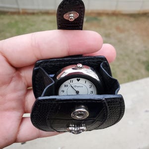 May include: A black Brighton watch with a white face and silver-tone accents, nestled in a matching black leather case. The watch features a heart symbol above the 6 and the Brighton logo. The case has a snap closure.