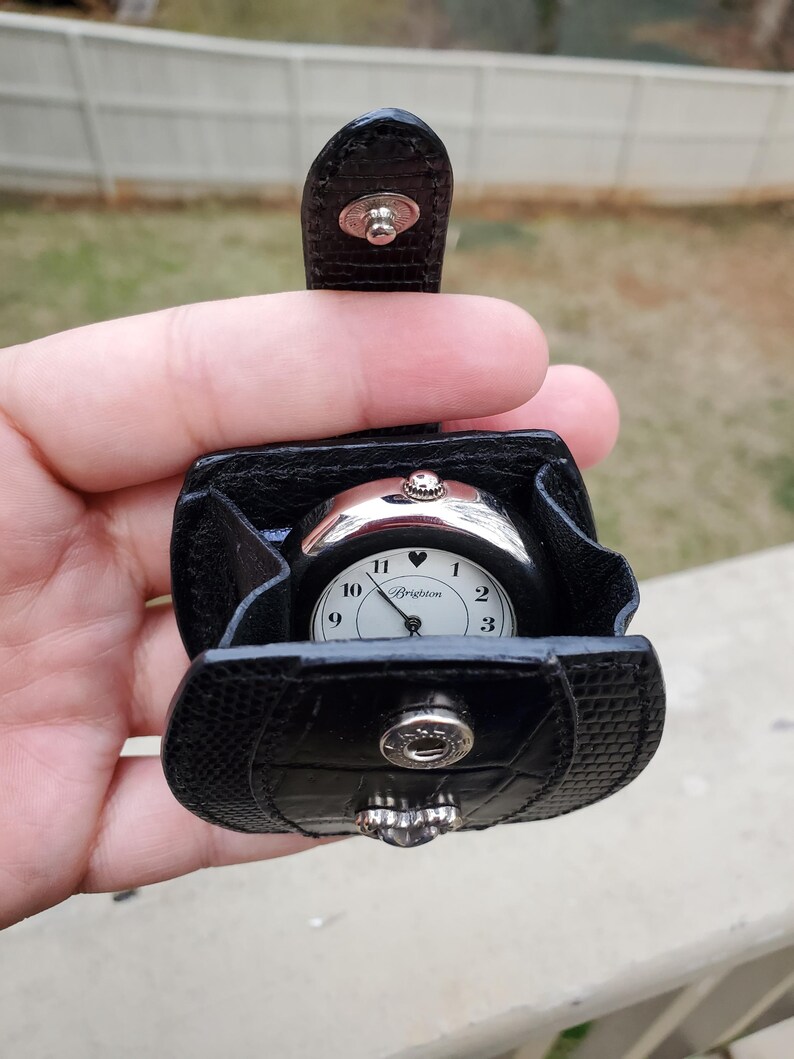 May include: A black Brighton watch with a white face and silver-tone accents, nestled in a matching black leather case. The watch features a heart symbol above the 6 and the Brighton logo. The case has a snap closure.