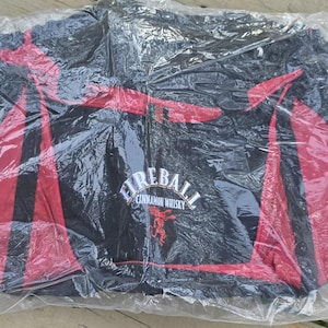 K&ouml;nnte beinhalten: Schwarze Reisetasche mit roten Akzenten, verpackt in durchsichtigem Kunststoff. Die Tasche zeigt das "Fireball Cinnamon Whisky"-Logo in Wei&szlig;, mit einem roten Drachenemblem. Geeignet f&uuml;r Reisen oder zum Tragen von Gegenst&auml;nden.