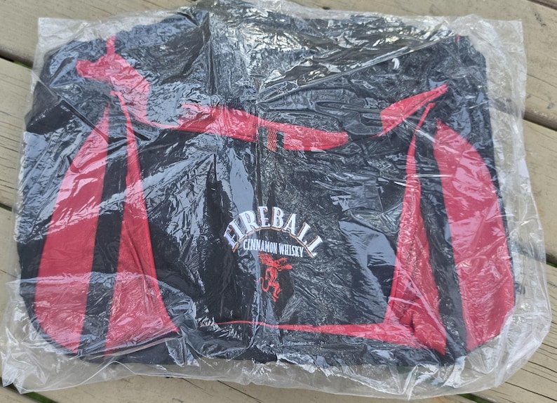 K&ouml;nnte beinhalten: Schwarze Reisetasche mit roten Akzenten, verpackt in durchsichtigem Kunststoff. Die Tasche zeigt das "Fireball Cinnamon Whisky"-Logo in Wei&szlig;, mit einem roten Drachenemblem. Geeignet f&uuml;r Reisen oder zum Tragen von Gegenst&auml;nden.