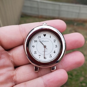 May include: A round, silver-toned Brighton quartz watch face with a white dial. Black numerals mark the hours, with a heart symbol at the 12 o'clock position. The watch has a small crown and two lugs for attachment.