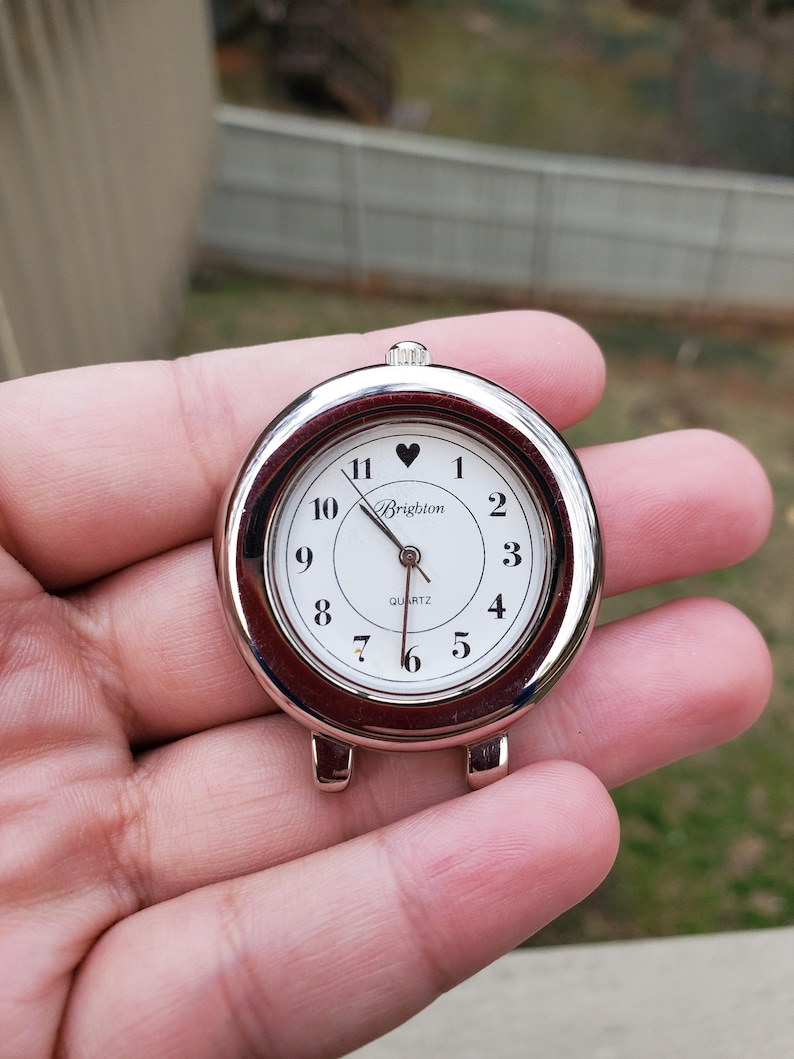 May include: A round, silver-toned Brighton quartz watch face with a white dial. Black numerals mark the hours, with a heart symbol at the 12 o'clock position. The watch has a small crown and two lugs for attachment.