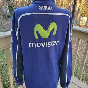 May include: A blue Yamaha Movistar zip-up sweatshirt. The back features a large lime green "M" logo above the word "Movistar" in white. White and black accents run down the sleeves. The Yamaha logo is at the top.
