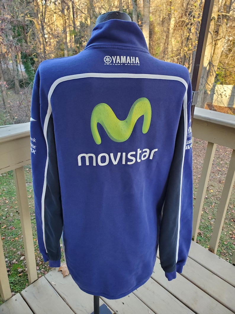 May include: A blue Yamaha Movistar zip-up sweatshirt. The back features a large lime green "M" logo above the word "Movistar" in white. White and black accents run down the sleeves. The Yamaha logo is at the top.