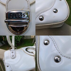Op de afbeelding: Een witte handtas met een zilverkleurige metalen sluiting met de in reli&euml;f aangebrachte tekst GUESS. De tas is voorzien van zilverkleurige ronde hardware, decoratieve metalen studs op de hoeken en witte stiksels op het buitenoppervlak.
