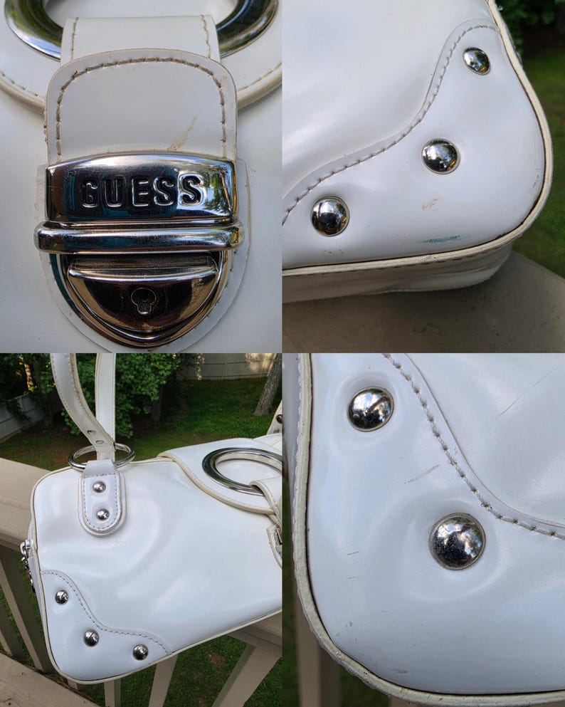 Op de afbeelding: Een witte handtas met een zilverkleurige metalen sluiting met de in reli&euml;f aangebrachte tekst GUESS. De tas is voorzien van zilverkleurige ronde hardware, decoratieve metalen studs op de hoeken en witte stiksels op het buitenoppervlak.