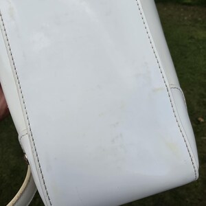 Op de afbeelding: Een witte lakleren handtas met een gladde, glanzende afwerking en zichtbare stiksels langs de randen. De tas heeft een gestructureerde rechthoekige basis en een gebogen handvat aan de zijkant, met tekenen van lichte verkleuring op het oppervlak.