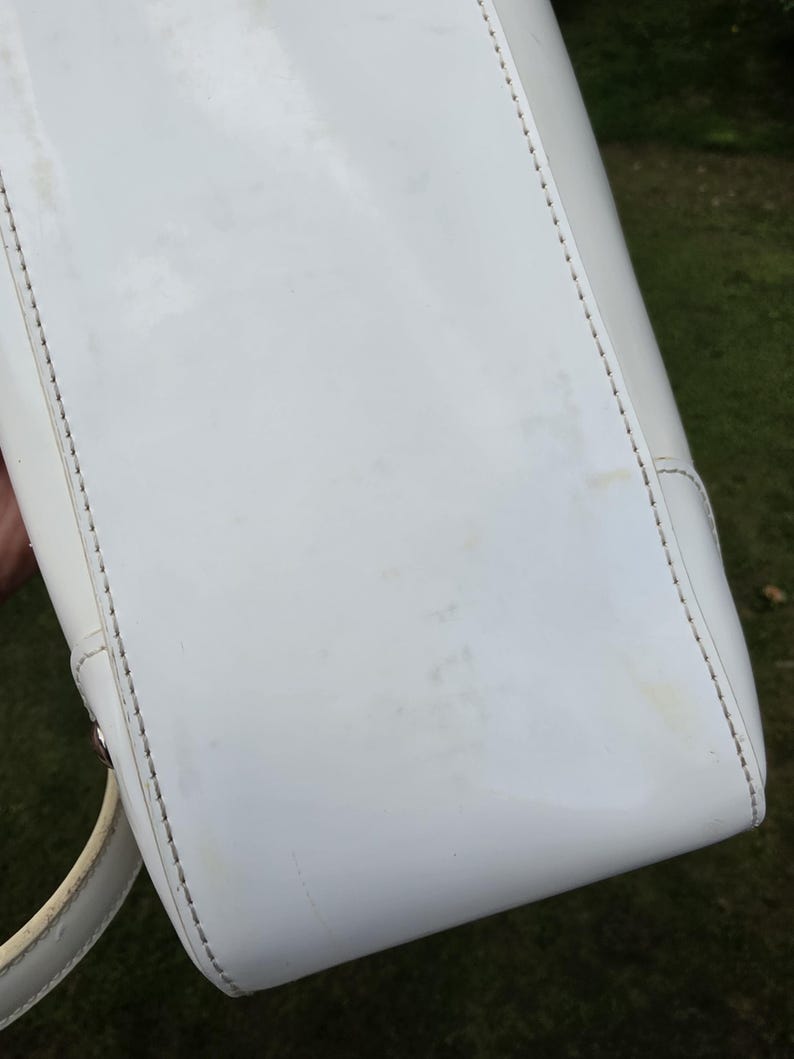 Op de afbeelding: Een witte lakleren handtas met een gladde, glanzende afwerking en zichtbare stiksels langs de randen. De tas heeft een gestructureerde rechthoekige basis en een gebogen handvat aan de zijkant, met tekenen van lichte verkleuring op het oppervlak.