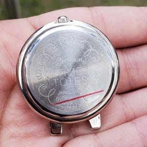 May include: A silver-toned watch back with the text "Brighton TIME TOTES JAPAN MOVT" and decorative swirls. The watch back has two small lugs and a crown. A red line is visible near the bottom.