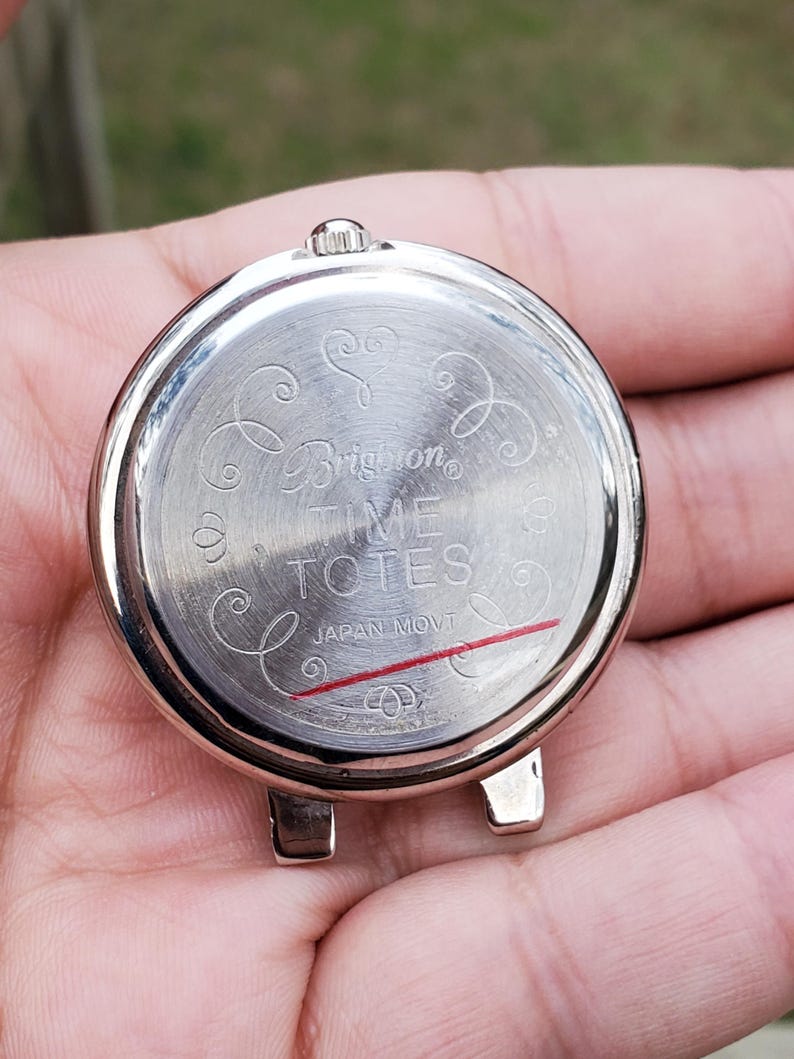 May include: A silver-toned watch back with the text "Brighton TIME TOTES JAPAN MOVT" and decorative swirls. The watch back has two small lugs and a crown. A red line is visible near the bottom.