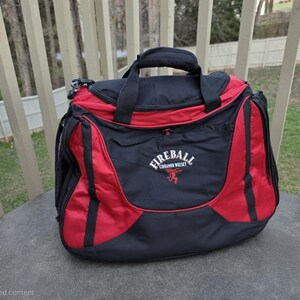 K&ouml;nnte beinhalten: Eine schwarz-rote Reisetasche mit oberem Griff und Schultergurtbefestigungen. Die Tasche tr&auml;gt das Logo "Fireball Cinnamon Whisky" in Wei&szlig; und eine rote Grafik. Die Tasche steht auf einer grauen Oberfl&auml;che.