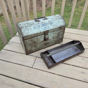 May include: A vintage metal toolbox in a distressed light blue-green finish, with the text "G.S. BUTLER 253". The toolbox has a handle and a latch. A metal tray is placed beside the toolbox, likely for tools.