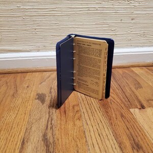 May include: A blue leather bound notebook with a ring binder. The notebook is open to a page with the title "Selected E. W. Co. Products" and a list of products with their prices. The notebook is sitting on a wooden floor.