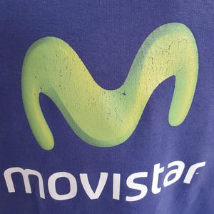 May include: A navy blue garment featuring a large, faded green logo in the shape of a stylized 'M'. Below the logo, the word "movistar" is printed in white, with a slightly distressed appearance.