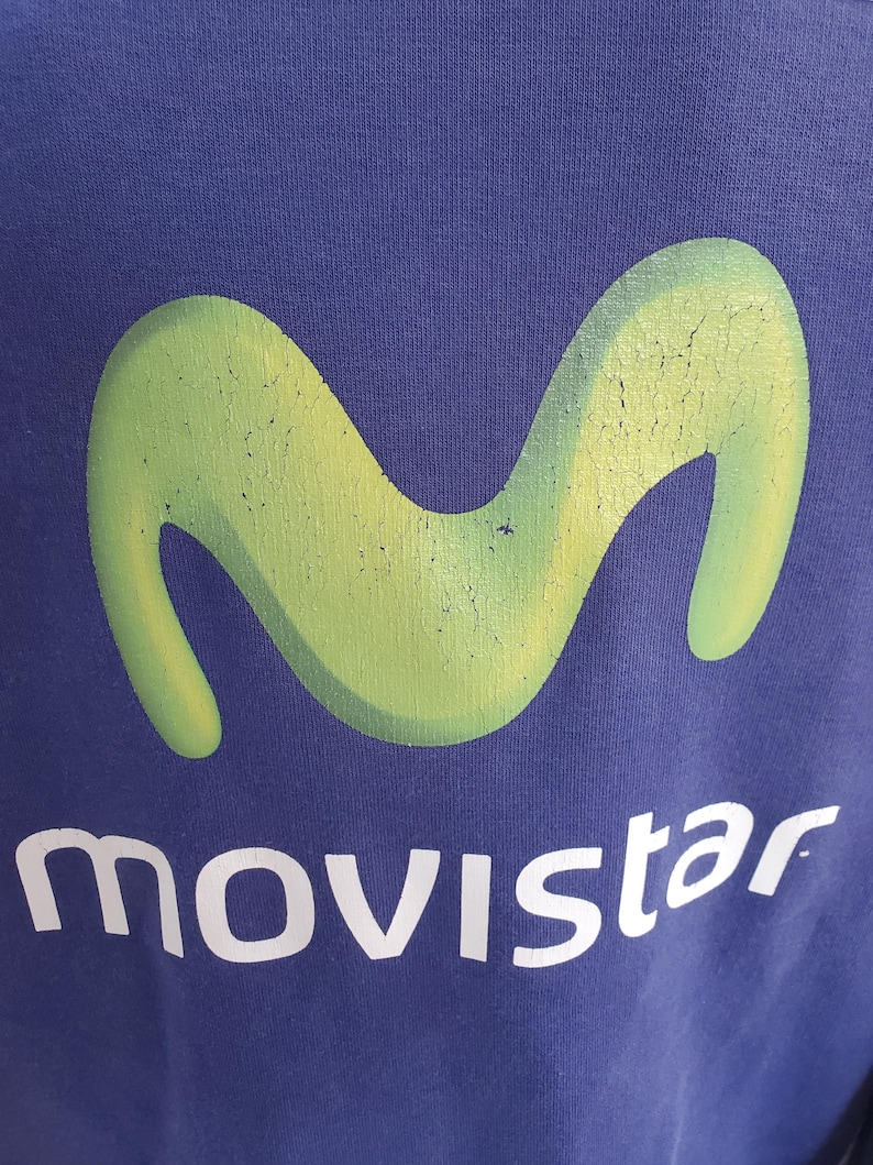 May include: A navy blue garment featuring a large, faded green logo in the shape of a stylized 'M'. Below the logo, the word "movistar" is printed in white, with a slightly distressed appearance.
