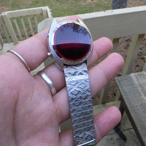 May include: A vintage silver-toned wristwatch with a unique design. The watch face is a deep red color, and the band is made of textured metal. The watch is held in a hand, showcasing its retro style and design.
