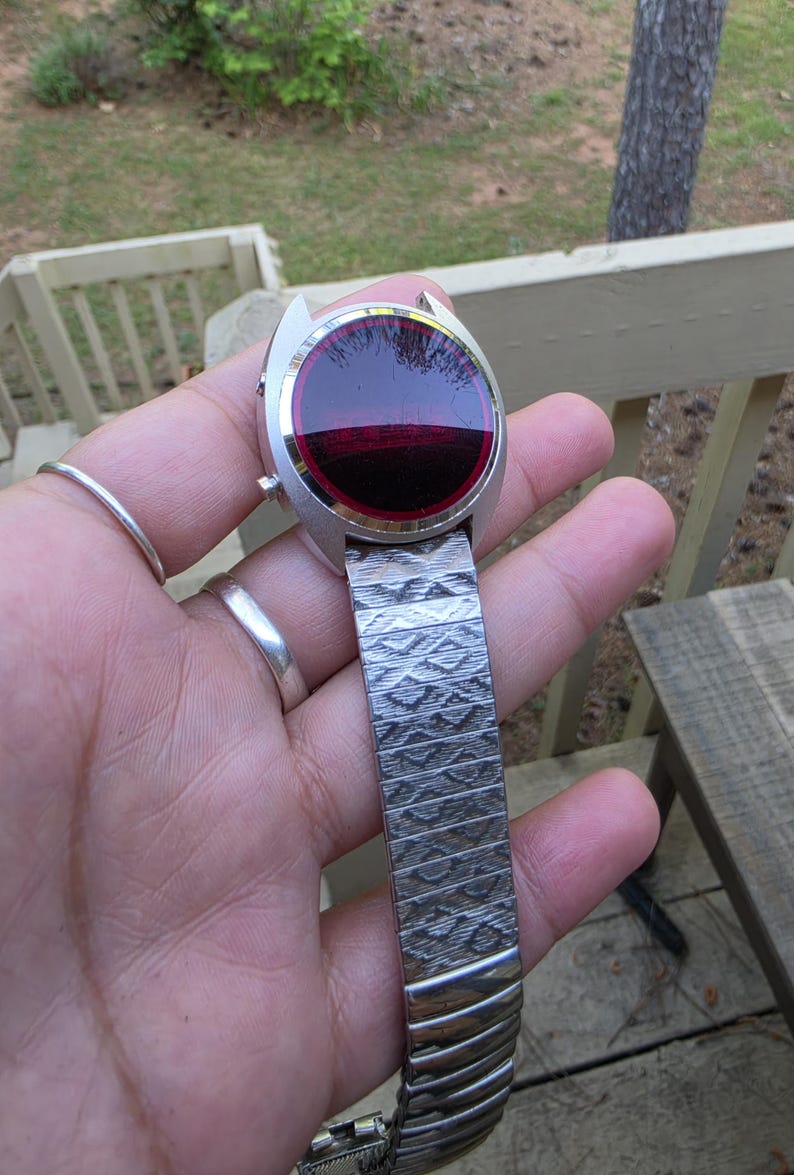 May include: A vintage silver-toned wristwatch with a unique design. The watch face is a deep red color, and the band is made of textured metal. The watch is held in a hand, showcasing its retro style and design.