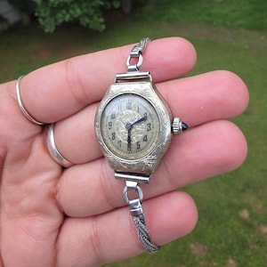 Pode incluir: Um relógio de pulso prateado com uma caixa de metal gravada e ornamentada, e um mostrador texturizado com algarismos arábicos pretos. O relógio inclui uma delicada pulseira de corrente metálica trançada e uma pequena pedra azul na coroa.