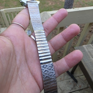 May include: A silver-toned wristwatch with a patterned metal band. The watch face is rectangular with a dark face. The band is made of interlocking metal links with a textured design. The watch is held up against a natural background.