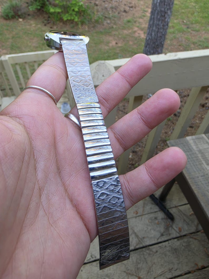 May include: A silver-toned wristwatch with a patterned metal band. The watch face is rectangular with a dark face. The band is made of interlocking metal links with a textured design. The watch is held up against a natural background.