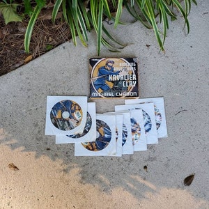 May include: A set of seven white discs with blue and gold designs, featuring the text "The Amazing Adventures of Kavalier & Clay" by Michael Chabon. The discs are arranged in a semi-circle on a concrete surface.