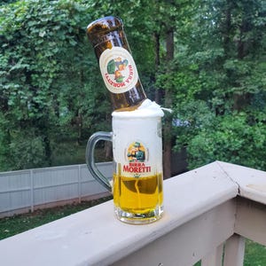 May include: A glass beer mug filled with golden beer and a thick white head, with a tilted brown beer bottle balanced on top. The mug features the "Birra Moretti" logo. The background shows a green, natural setting.