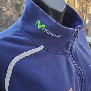 May include: A navy blue zip-up jacket with a high collar. The jacket features a white reflective stripe along the shoulder and the Movistar logo in green and white. An orange and white Eneos logo is visible on the lower right.