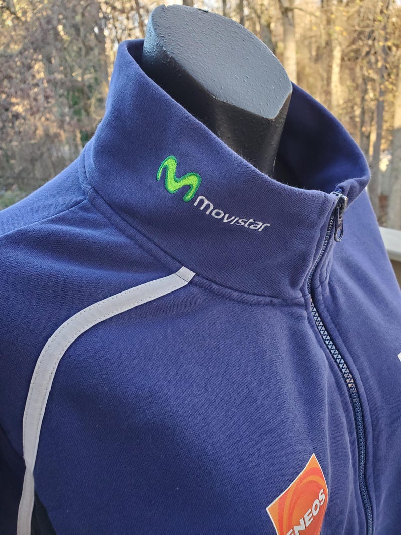 May include: A navy blue zip-up jacket with a high collar. The jacket features a white reflective stripe along the shoulder and the Movistar logo in green and white. An orange and white Eneos logo is visible on the lower right.