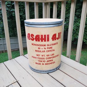 May include: A cylindrical container of "ASAHI AJI" monosodium glutamate. The container is beige with red lettering, including the text "MONOSODIUM GLUTAMATE 99 + % PURE REGULAR CRYSTAL" and "NET WT. 100 LBS". The container has metal bands and a lid.