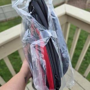 K&ouml;nnte beinhalten: Eine schwarze Rei&szlig;verschlusstasche mit rotem Streifen, verpackt in durchsichtigem Kunststoff. Die Tasche scheint gefaltet und mit einem Kunststoffband befestigt zu sein. Der Hintergrund zeigt ein Holzdeck und Gr&uuml;n.