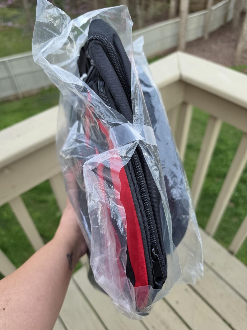 K&ouml;nnte beinhalten: Eine schwarze Rei&szlig;verschlusstasche mit rotem Streifen, verpackt in durchsichtigem Kunststoff. Die Tasche scheint gefaltet und mit einem Kunststoffband befestigt zu sein. Der Hintergrund zeigt ein Holzdeck und Gr&uuml;n.
