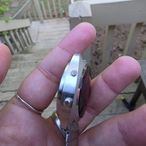 May include: A silver-toned wristwatch with a rectangular face and a metal band. The watch has a dark-colored display and two small buttons on the side. The band is made of small, connected metal pieces.