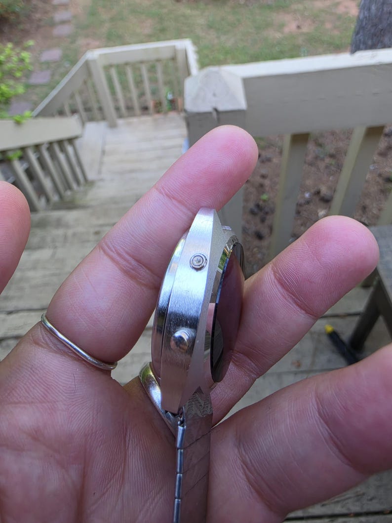 May include: A silver-toned wristwatch with a rectangular face and a metal band. The watch has a dark-colored display and two small buttons on the side. The band is made of small, connected metal pieces.
