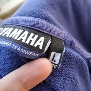 May include: Close-up of a blue Yamaha Teamwear sweatshirt tag. The tag is black with white text, and the size is indicated as Large (L). The sweatshirt is a solid blue color.