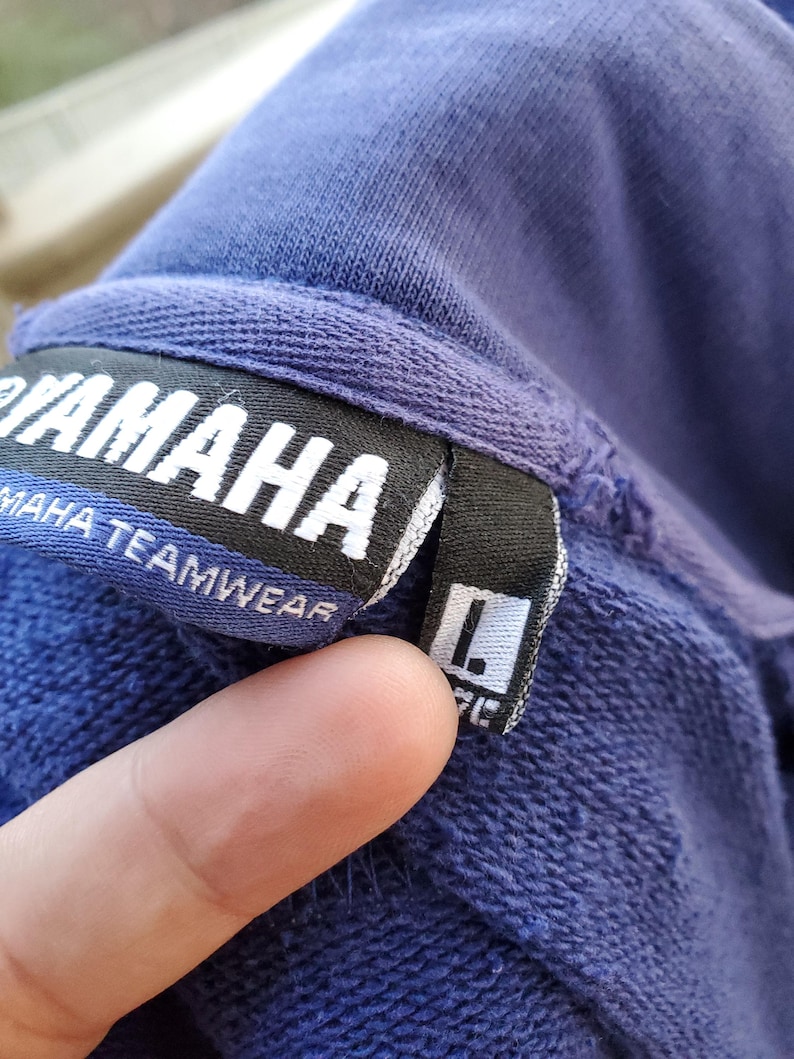 May include: Close-up of a blue Yamaha Teamwear sweatshirt tag. The tag is black with white text, and the size is indicated as Large (L). The sweatshirt is a solid blue color.