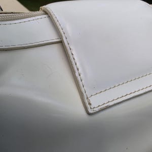 Op de afbeelding: Een witte handtas met een gladde, glanzende afwerking en een opvallende klepsluiting. De randen van de klep zijn verstevigd met zichtbare witte stiksels. Een rits loopt langs de bovenrand en een metalen ring is zichtbaar aan de hoek.