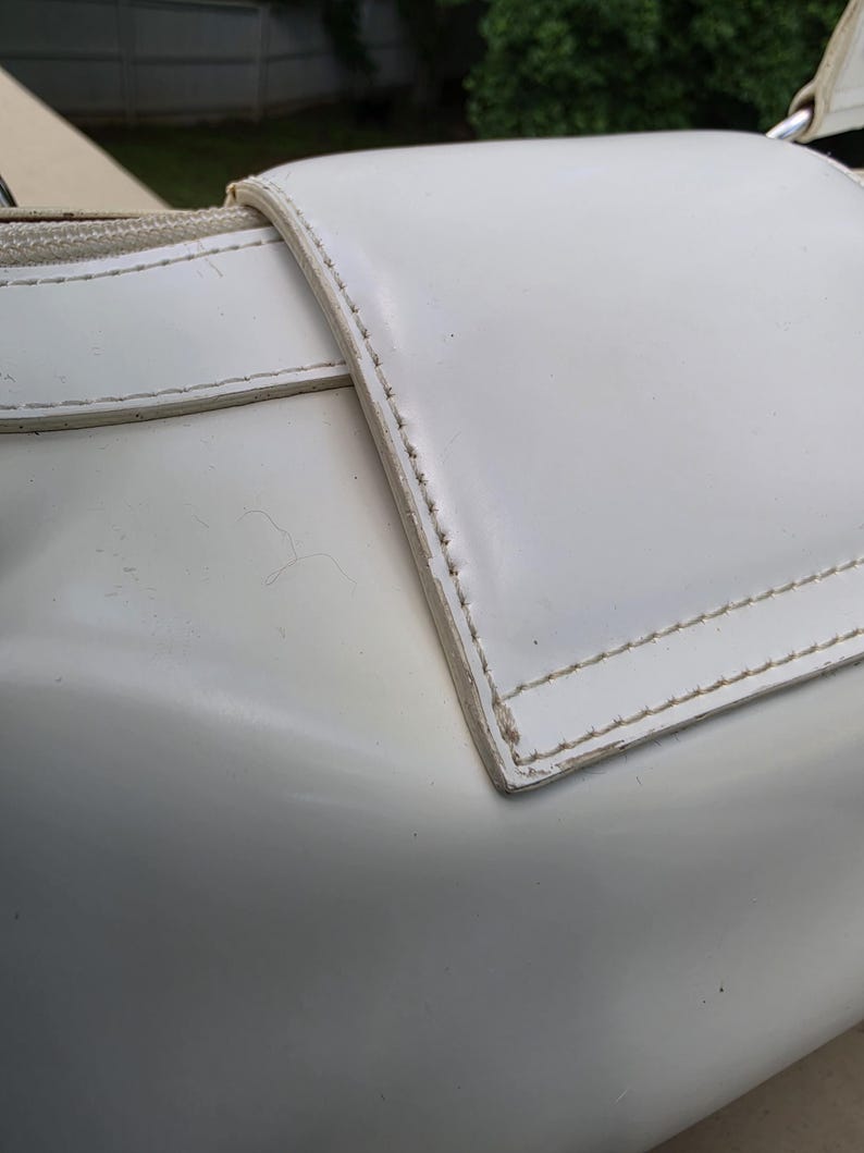 Op de afbeelding: Een witte handtas met een gladde, glanzende afwerking en een opvallende klepsluiting. De randen van de klep zijn verstevigd met zichtbare witte stiksels. Een rits loopt langs de bovenrand en een metalen ring is zichtbaar aan de hoek.