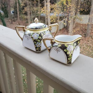 May include: A vintage tea set featuring a teapot with a lid and a creamer. The set is white with gold trim, black and yellow accents, and floral designs. The teapot has a gold knob and handle. The creamer has a matching design.