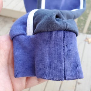 May include: Close-up of a blue sweatshirt sleeve with a ribbed cuff. The sleeve has a white stripe and a dark gray stripe running down the side. The cuff is a darker shade of blue.