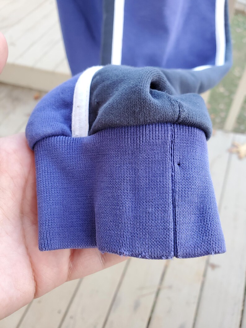 May include: Close-up of a blue sweatshirt sleeve with a ribbed cuff. The sleeve has a white stripe and a dark gray stripe running down the side. The cuff is a darker shade of blue.