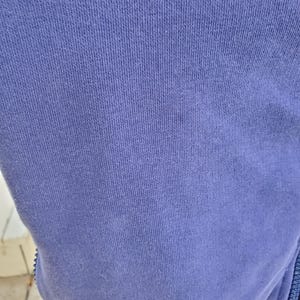 May include: Close-up of a section of a periwinkle blue fabric. The fabric has a ribbed texture, with vertical lines running across the surface. The material appears to be a soft knit, possibly cotton or a cotton blend. A zipper is visible on the right side.