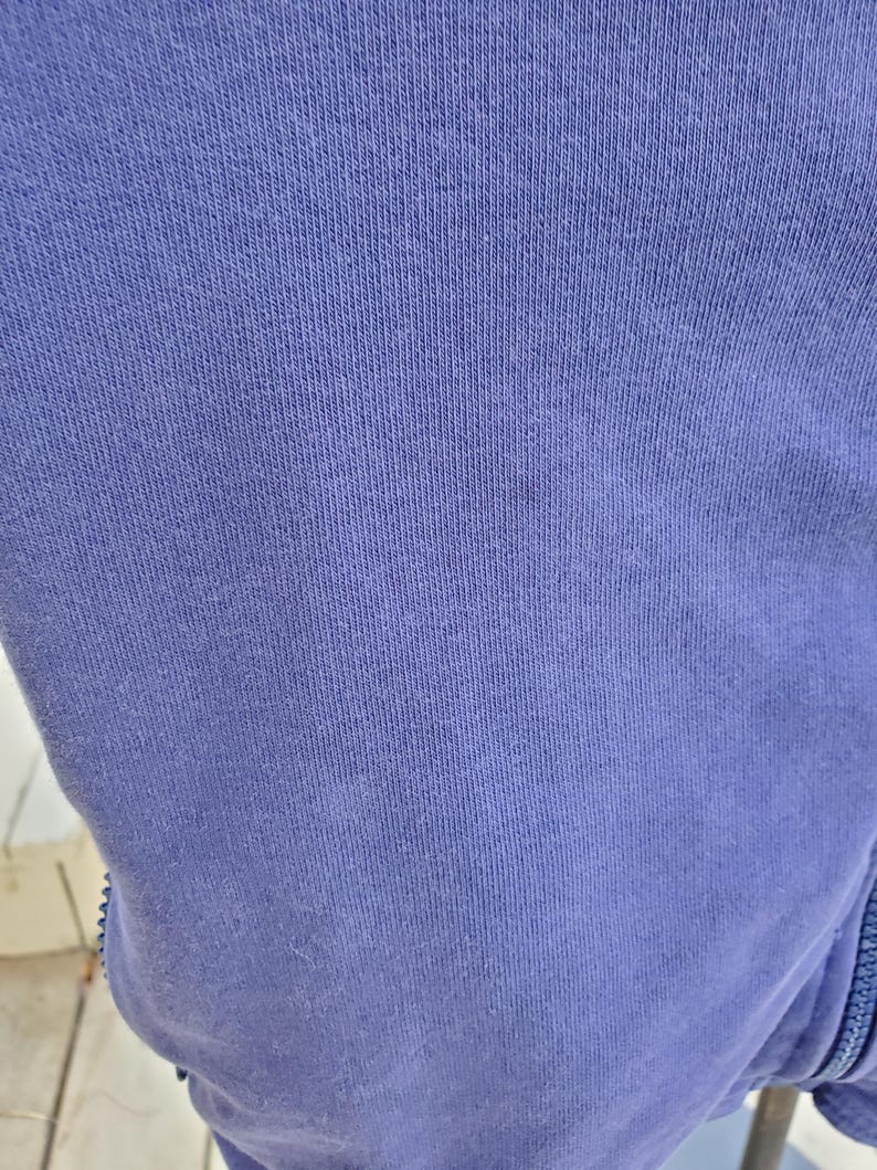 May include: Close-up of a section of a periwinkle blue fabric. The fabric has a ribbed texture, with vertical lines running across the surface. The material appears to be a soft knit, possibly cotton or a cotton blend. A zipper is visible on the right side.