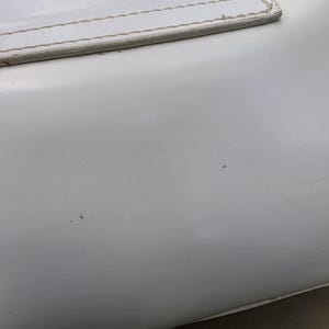 Op de afbeelding: Een glad, glanzend wit oppervlak met een rechthoekig paneel met zichtbare stiksels langs de bovenrand. Het materiaal lijkt een synthetische of leerachtige afwerking te hebben, met een schone, effen witte kleur en subtiele reflecties.