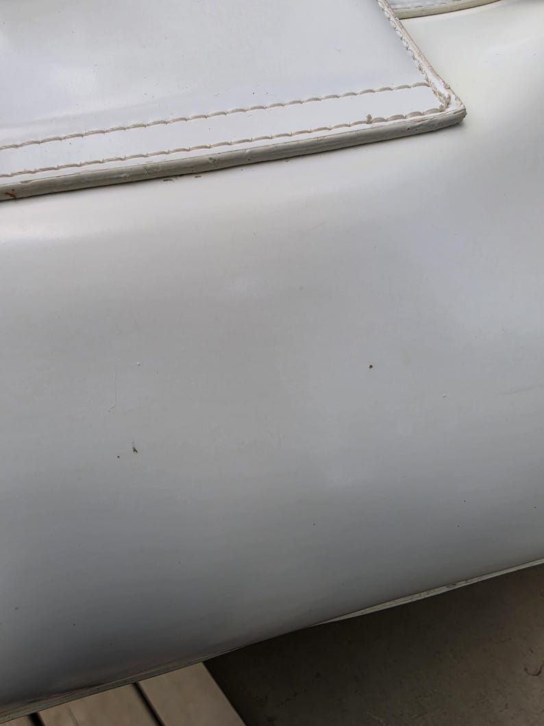 Op de afbeelding: Een glad, glanzend wit oppervlak met een rechthoekig paneel met zichtbare stiksels langs de bovenrand. Het materiaal lijkt een synthetische of leerachtige afwerking te hebben, met een schone, effen witte kleur en subtiele reflecties.