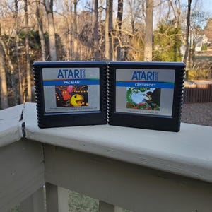 May include: Two black Atari video game cartridges. The left cartridge has a label that reads "ATARI PAC-MAN". The right cartridge has a label that reads "ATARI CENTIPEDE".
