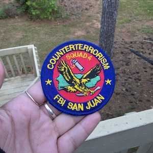 May include: A circular patch with the words "COUNTERTERRORISM SQUAD 4" in yellow and red. The patch features a yellow eagle with black wings, a map, and the text "FBI SAN JUAN". The patch is blue with yellow stars.