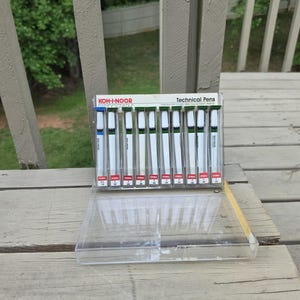 May include: A package of Koh-I-Noor technical pens. The pens are white with black and blue accents, displayed in a clear plastic case. The packaging includes the text "KOH-I-NOOR" and "Technical Pens". The pens are on a wooden surface.